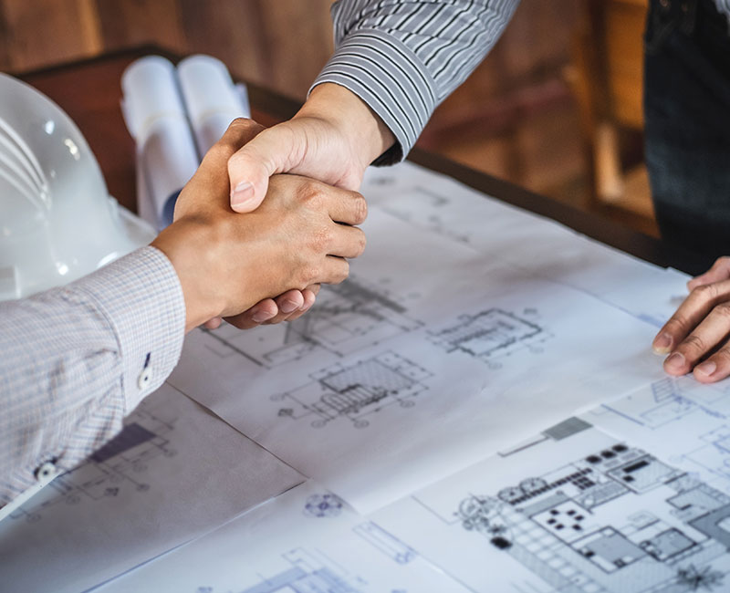 Two people shaking hands over architectural plans, with a white hard hat and blueprints nearby, set in a warm, wooden environment.