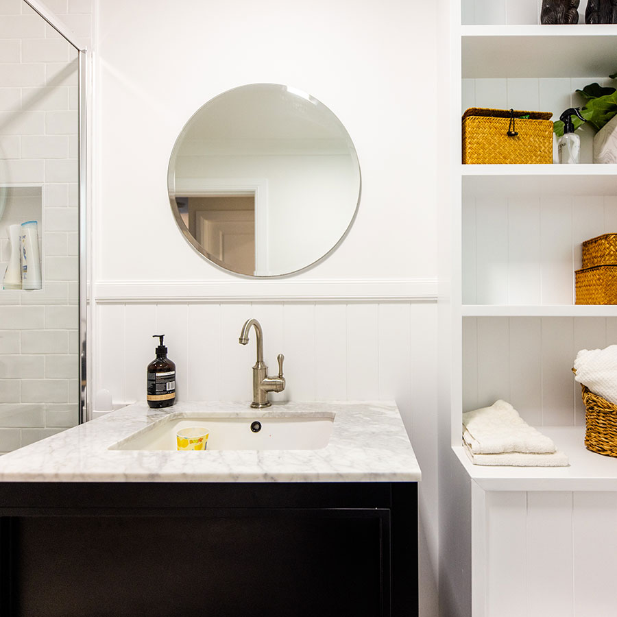Modern bathroom with a round mirror, marble sink, and black cabinetry; white shelves display woven baskets and towels.