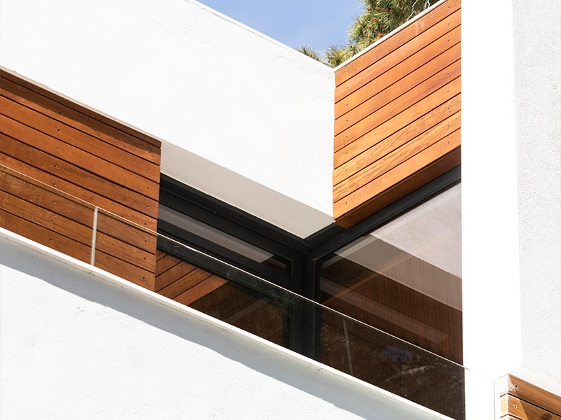 Modern architectural design featuring a white exterior with wooden panel accents, large glass windows, and a bright blue sky in the background.