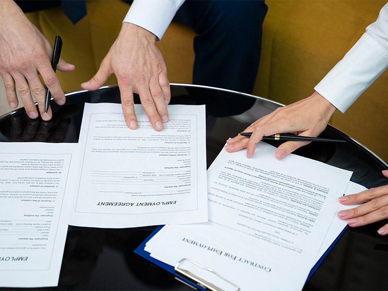 Hands of two individuals sign various documents on a glossy table, with white papers arranged around a blue folder.