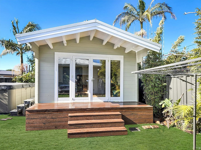 Charming garden shed featuring large windows, a sloped roof, and wooden steps, surrounded by lush greenery and palm trees.