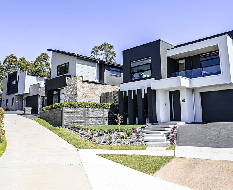Modern residential buildings featuring a mix of dark and light colours, landscaped gardens, and driveways on a gentle slope.