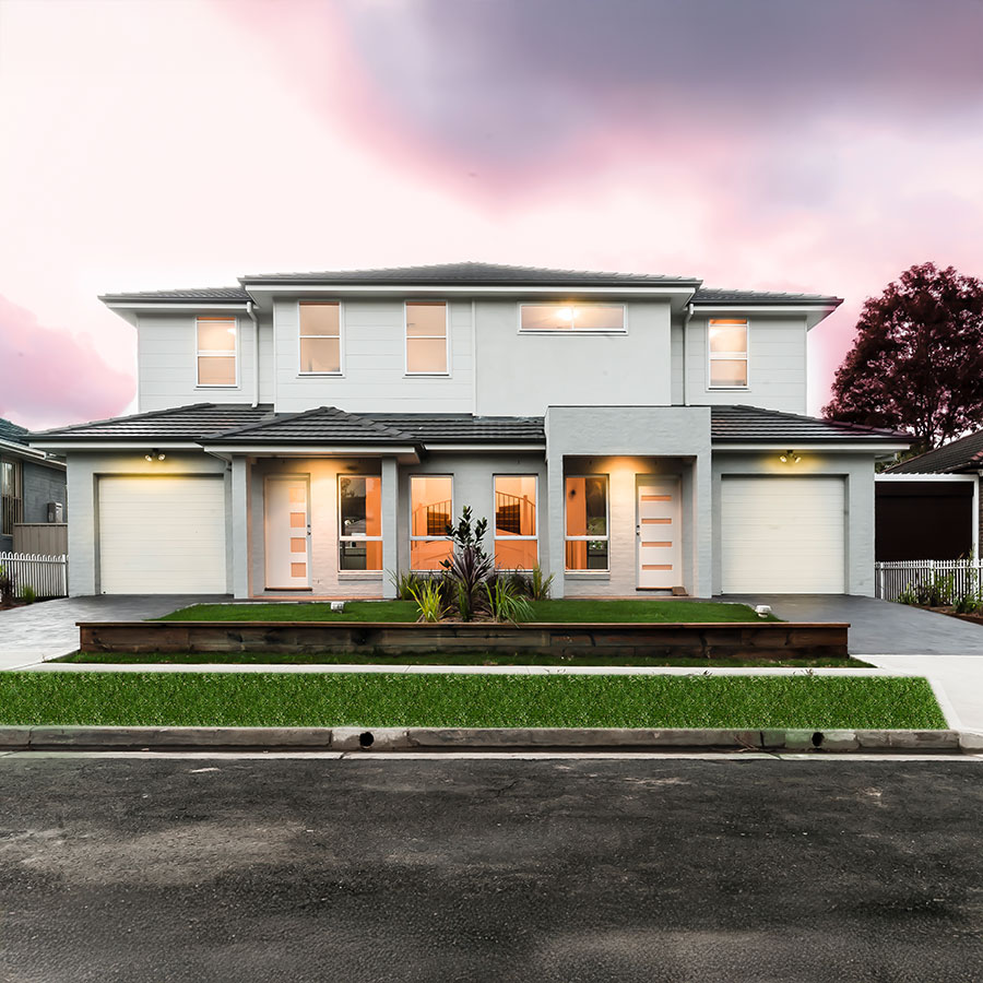Modern duplex with a light grey façade, stone features, and landscaped front garden, set against a pink-tinted sky at dusk.