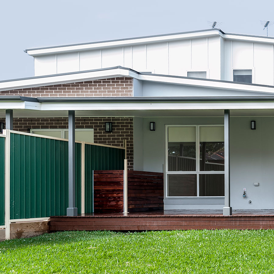 Modern duplex featuring a blend of brick and weatherboard exterior, with a green fence, wooden deck, and landscaped lawn.