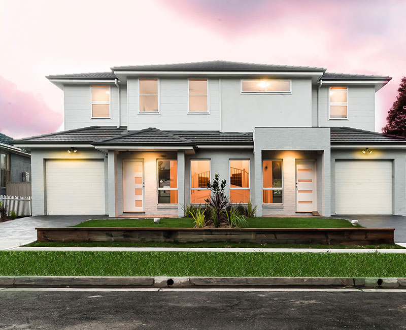 A modern duplex featuring two symmetrical units, light grey exterior, large windows, and a landscaped front garden with greenery.