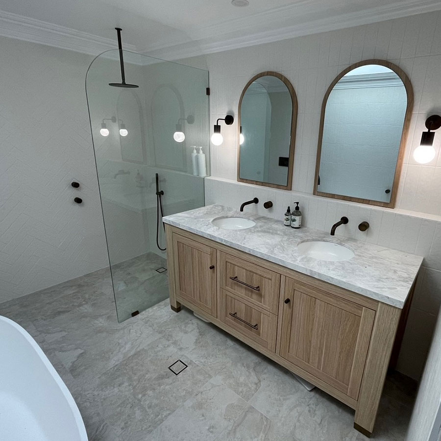 Modern bathroom featuring a wooden double vanity with marble top, two mirrors, a glass shower, and neutral-toned tiles.