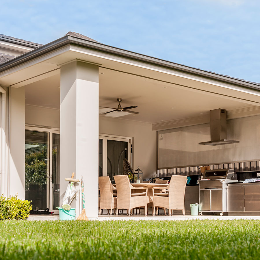 Modern outdoor dining space with a large table and wicker chairs, featuring a barbecue area and lush green grass. Clear blue sky above.