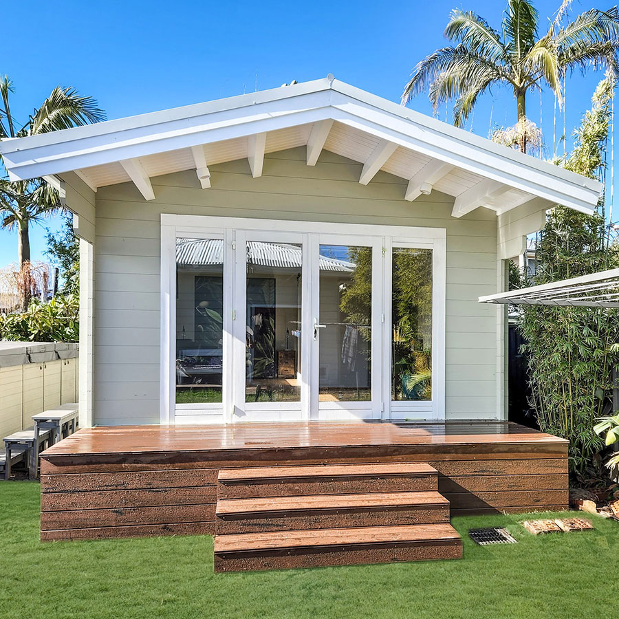 A modern light-green house with a sloped white roof, featuring large glass sliding doors and wooden steps on a grassy area, surrounded by palm trees.