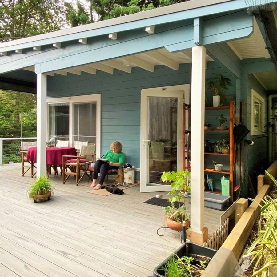 A light blue granny flat features a covered porch with wooden decking. A woman sits on a chair, surrounded by potted plants and outdoor furniture.