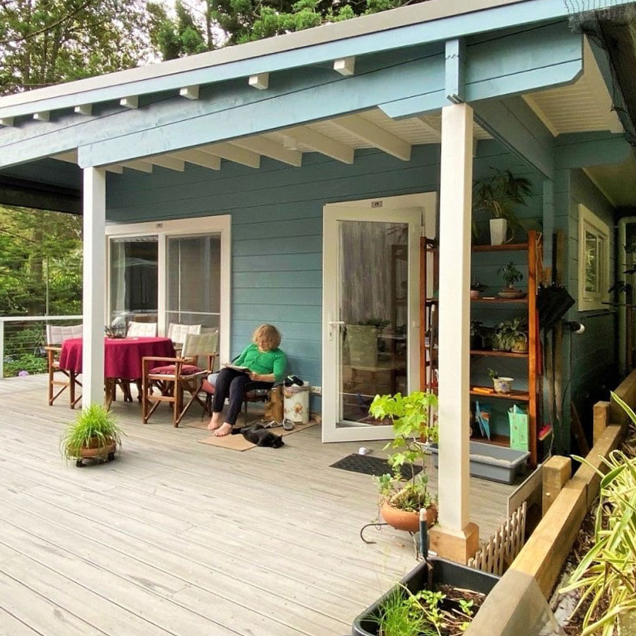 A woman sits on a chair on a wooden deck outside a blue house, with a table set for dining and various plants around.