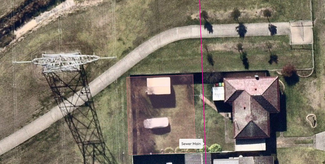 Aerial view of a residential area showing a house and adjacent property with a marked sewer main, surrounded by green lawns and pathways.